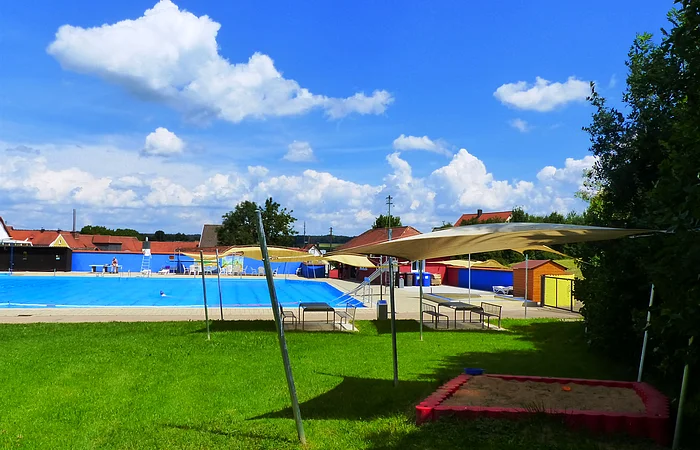 Ein Freibad mit Schwimmbecken, Sonnensegeln, Sitzmöglichkeiten und einem Sandkasten auf einer grünen Wiese, rechts unten im Bild. Im Hintergrund blauer Himmel mit Wolken.
