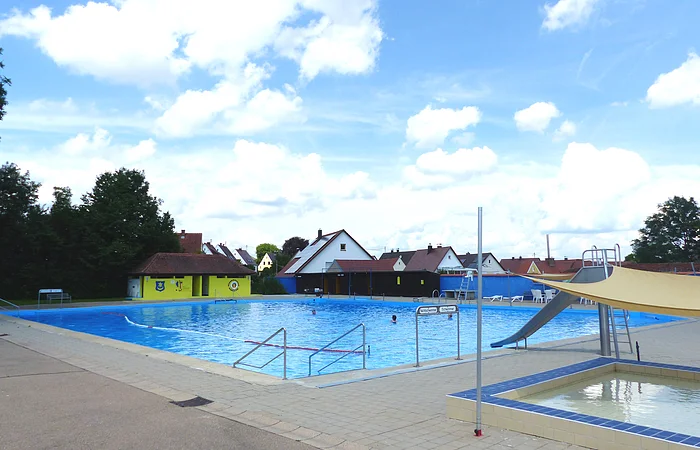 Ein Freibad mit einem Schwimmbecken, einer Rutsche und einem Kinderbecken. Im Hintergrund steht ein grünes Gebäude mit rotem Dach. Dahinter sind Wohnhäuser und Bäume und blauer Himmel zu sehen.