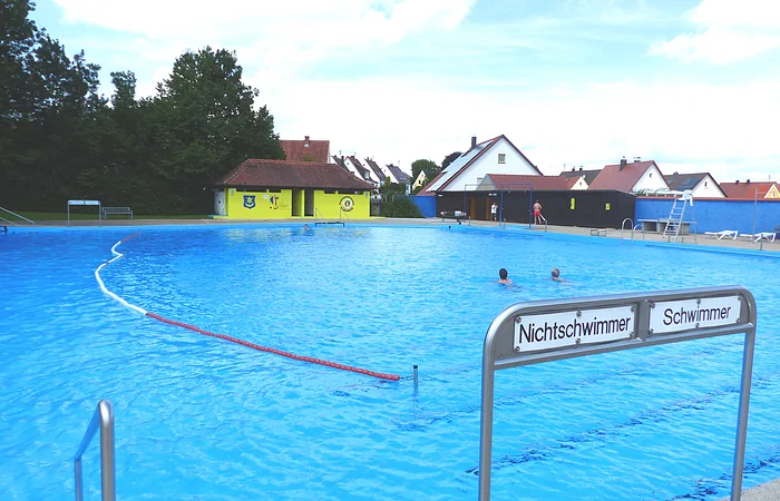 Ein Freibad mit einem großen Schwimmbecken, unterteilt in Nichtschwimmer- und Schwimmerbereich. Im Hintergrund sind Gebäude und Bäume zu sehen.