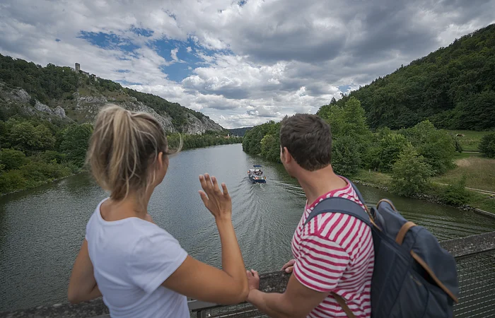 Eine Frau mit weißen Shirt und ein Mann mit Rucksack stehen mit dem Blick auf den Main-Donau-Kanal gerichtet auf der Holzbrücke am Geländer. Der Himmel ist sehr bedeckt. Die beiden Ufer erstrahlen im dunklen grün.
