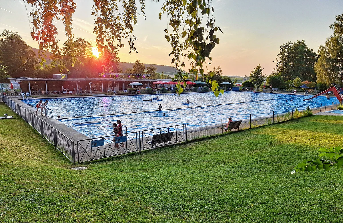 Das Freibad ist umgeben von grüner Wiese und Bäumen. Die Sonne ist gerade am Untergehen.