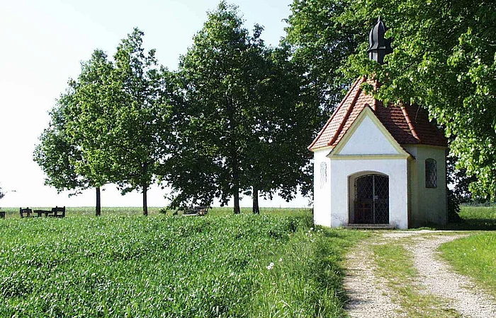Kleine Kapelle mit rotem Dach auf einer Wiese mit einem Schotterweg. Umgeben von einem grünem Feld und Bäumen.