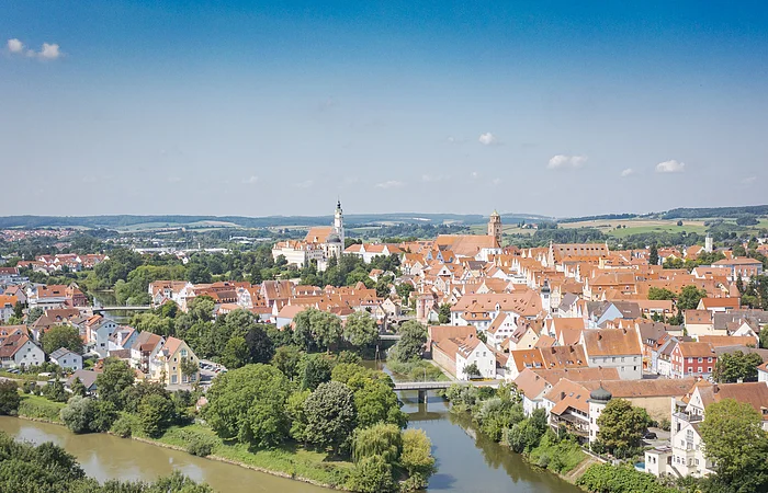Luftaufnahme einer Stadt mit roten Dächern, Fluss und Brücke, umgeben von grüner Landschaft.