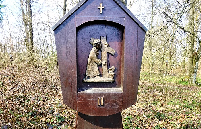 Kreuzwegstation bei der Brandkapelle in Monheim mit einer Holzstatue im Wald zeigt Jesus mit Kreuz. Oberhalb der Statue ein Kreuz, unterhalb die römische Nummer II. Hinter dem Wald bewölkter Himmel.