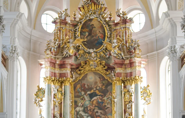 Zwei Personen mit Rucksäcken stehen in einer Kirche vor einem reich verzierten Altar.