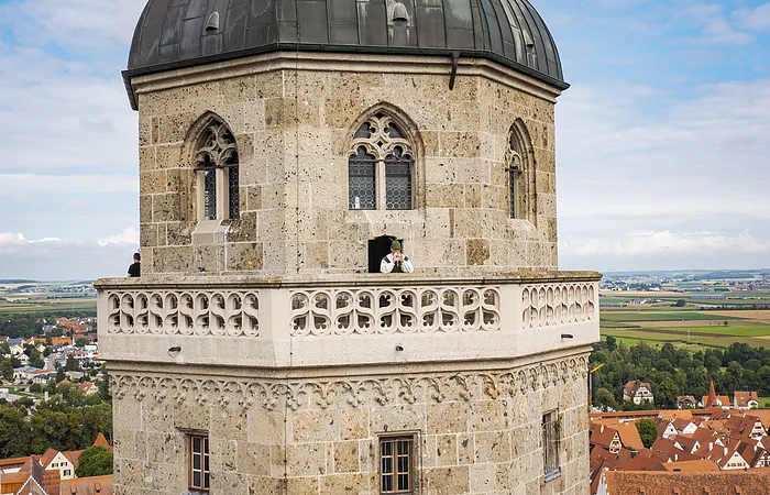 Steinerner Turm mit Rundbogenfenstern und Balustrade, Person mit Blasinstrument auf dem Balkon.