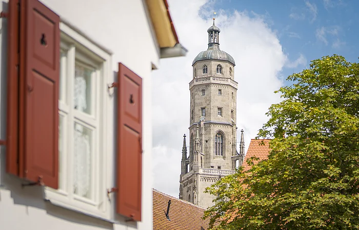 Ein Kirchturm ragt hinter einem Gebäude mit roten Fensterläden und einem Baum hervor.