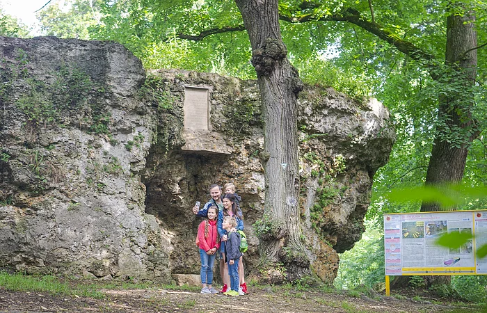 Eine Gruppe von Menschen steht vor einer großen Felsformation im Wald. Daneben befindet sich ein Informationsschild.