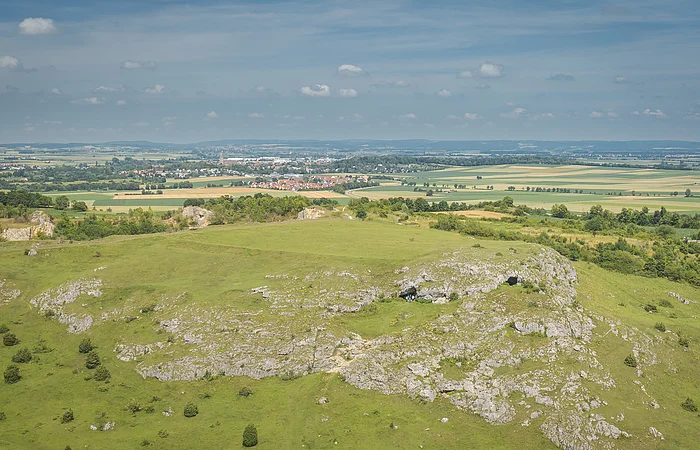 Hügelige Landschaft mit grünen Wiesen und Felsen im Vordergrund, Felder und ein Dorf im Hintergrund.
