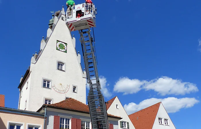 Feuerwehrleiterwagen mit ausgefahrener Leiter vor einem historischen Gebäude mit Torbogen und einem Turm dahinter. Auf dem Turm eine Sonnenuhr. Auf der Feuerwehrleiter stehen zwei Personen.