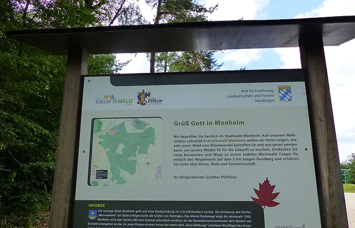 Ein überdachtes Informationsschild aus Holz im Wald mit Text über den Stadtwald Monheim und einer Karte des Waldgebiets.