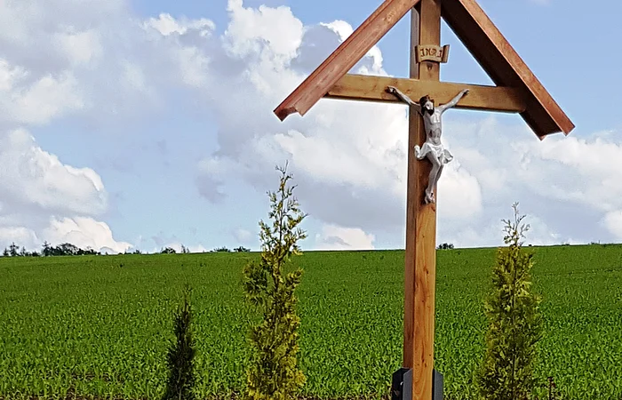 Holzkreuz mit überdachter Christusfigur mit bepflanztem Beet darunter, daneben eine Holzbank. Im Hintergrund ein Feld und blauer Himmel.