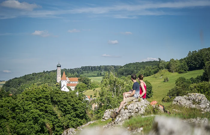 Zwei Personen sitzen auf einem Felsen und blicken auf eine Kirche in einer grünen Landschaft.