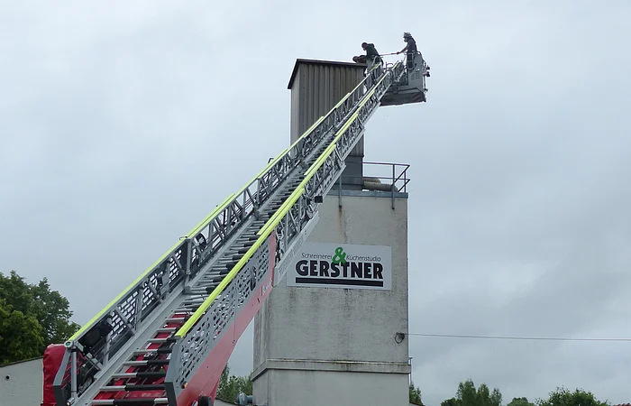 Feuerwehrwagen mit ausgefahrener Leiter vor einem Gebäude mit "Gerstner"-Schild. Auf der Leiter sind zwei Personen zu sehen, die Störche beringen. Im Hintergrund bewölkter Himmel.