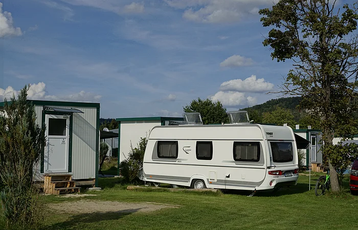 Wohnwagen auf einem Campingplatz neben kleinen Gebäuden und einem Baum.
