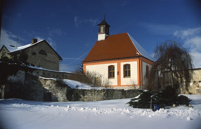 Verschneite Landschaft mit einer kleinen Kirche und einem angrenzenden Gebäude unter blauem Himmel.