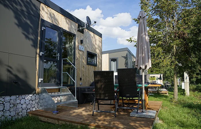 Moderne Holzhütte mit Terrasse, Tisch, Stühlen und Sonnenschirm im Freien, umgeben von Bäumen.