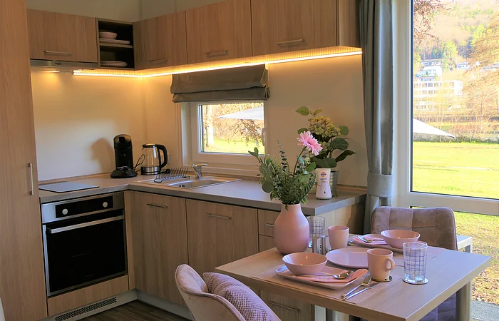 Kleine Küche mit Holzschränken, Elektrogeräten, Spüle und Esstisch. Fenster mit Blick ins Freie.