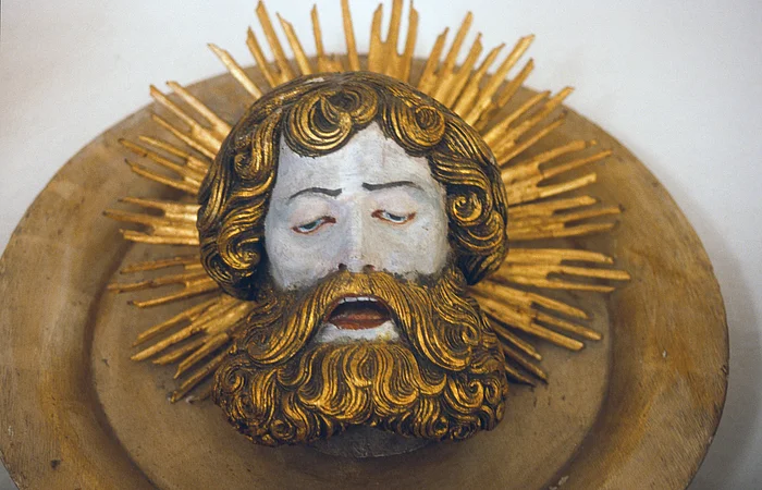 Holzskulptur eines bärtigen Männerkopfes mit geschlossenen Augen, umgeben von goldenen Strahlen.