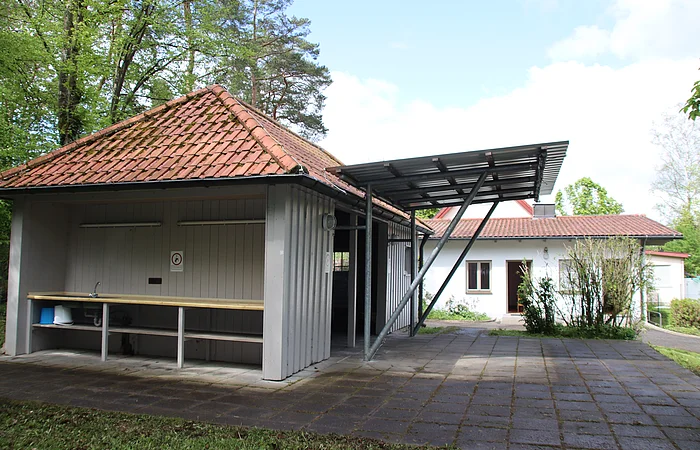 Ein kleiner, überdachter Waschplatz mit Spüle und Arbeitsfläche im Freien, umgeben von Bäumen.