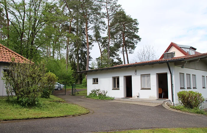 Ein einstöckiges weißes Gebäude mit mehreren Fenstern und offenen Türen, umgeben von Bäumen und einem gepflasterten Weg.