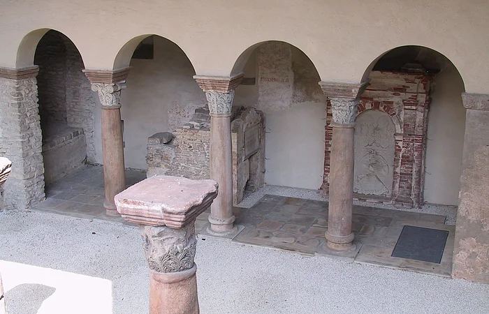 Steinarkaden mit verzierten Säulen in einem historischen Gebäude. Im Hintergrund sind Mauerreste sichtbar.