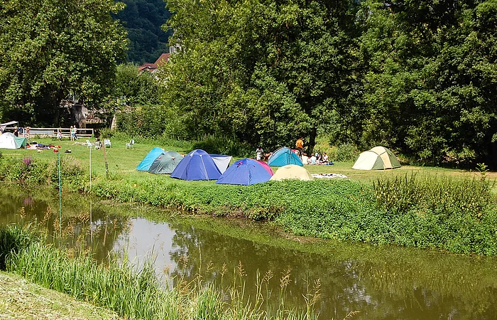 Zelte auf einer Wiese neben einem Fluss, umgeben von Bäumen und Menschen im Hintergrund.