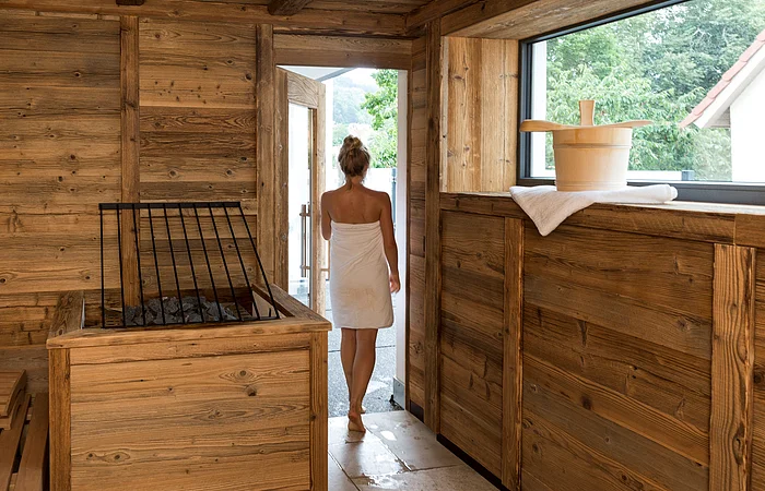 Eine Person in einem Handtuch verlässt eine Holzsauna durch eine Tür. Ein Saunaeimer steht auf einem Handtuch am Fenster.