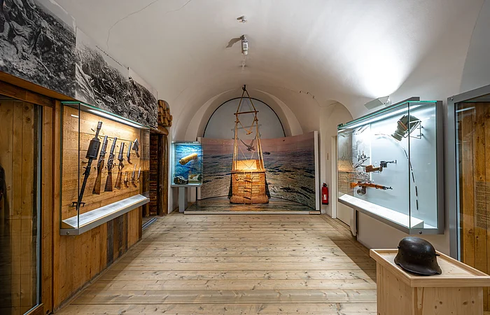 Ein Museumsraum mit Vitrinen, die historische Waffen und Ausrüstungsgegenstände zeigen. Ein Helm steht auf einem Holzpodest.