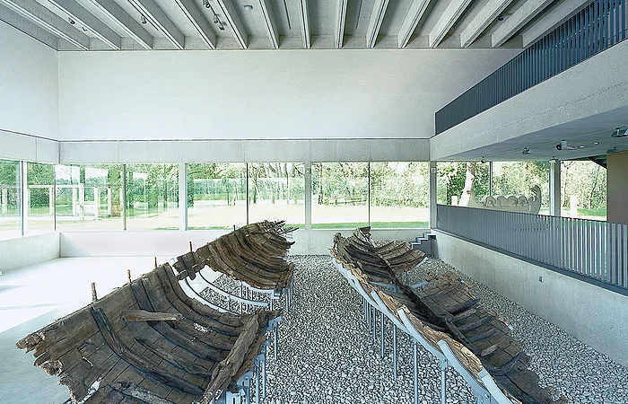 Zwei antike Holzbootwracks auf Kiesboden in einem modernen Ausstellungsraum mit Glaswänden.