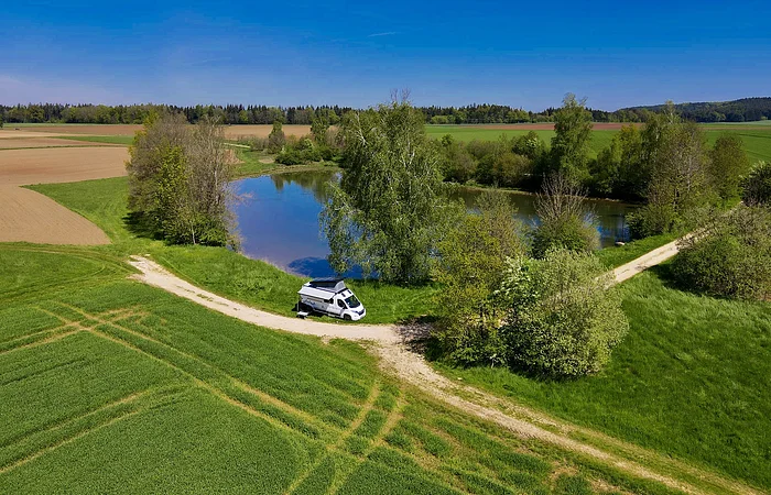 Wohnmobil auf einem Feldweg neben einem kleinen See, umgeben von Wiesen und Bäumen.