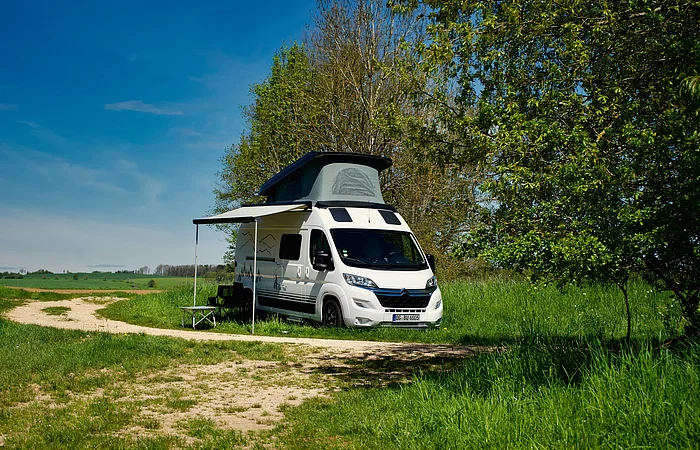 Wohnmobil mit ausgefahrenem Dach und Markise steht auf einer Wiese neben einem Baum.