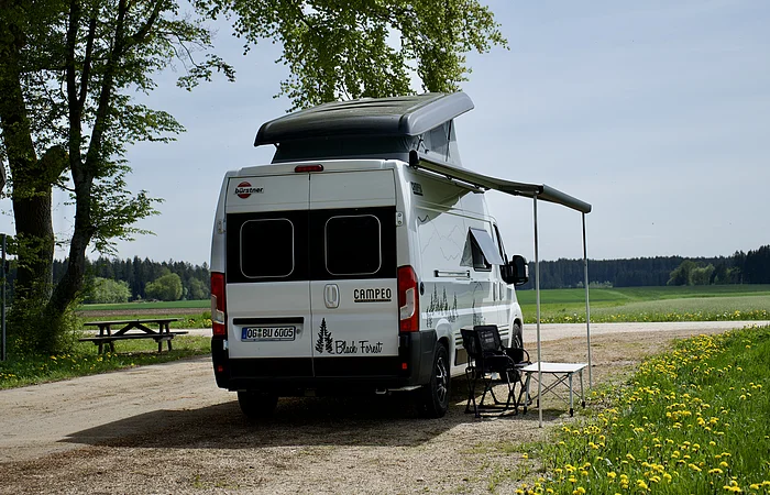 Wohnmobil mit ausgefahrener Markise steht auf einem Feldweg neben einer Wiese mit Löwenzahn.
