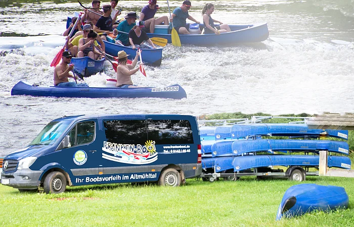 Eine Gruppe von Menschen paddelt in Kanus auf einem Fluss. Darunter steht ein Transporter mit Kanus auf einem Anhänger.