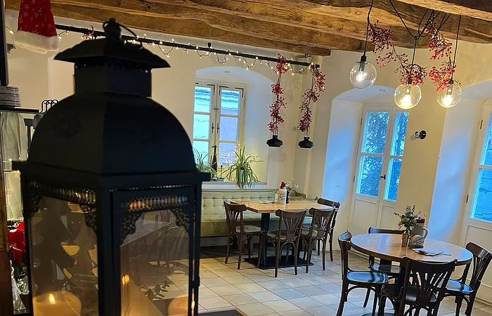 Gemütlich eingerichtetes Café mit Holzbalkendecke, Tischen und Stühlen. Eine Laterne mit Kerze steht im Vordergrund.