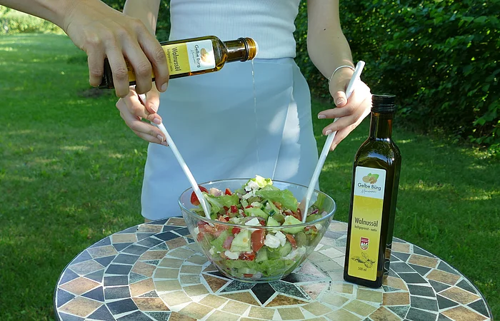 Person gießt Walnussöl in eine Schüssel mit Salat auf einem Mosaiktisch im Freien.