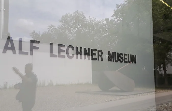 Glasfassade mit Schriftzug "Alf Lechner Museum", Spiegelung von Bäumen und einer Person.