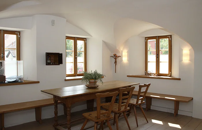 Esszimmer mit Holztisch, Stühlen und Bänken. Zwei Fenster, weiße Wände, Pflanze auf dem Tisch.