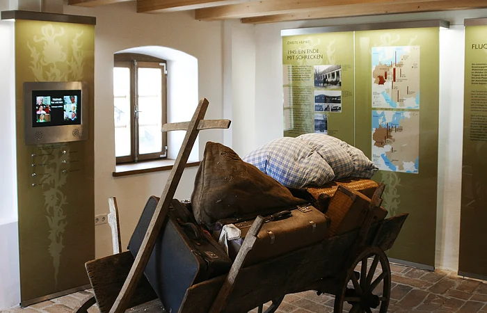 Ein Museumsausstellungsraum mit einer alten Holzkarren, beladen mit Gepäck und einem Holzkreuz. Im Hintergrund sind Informationstafeln.