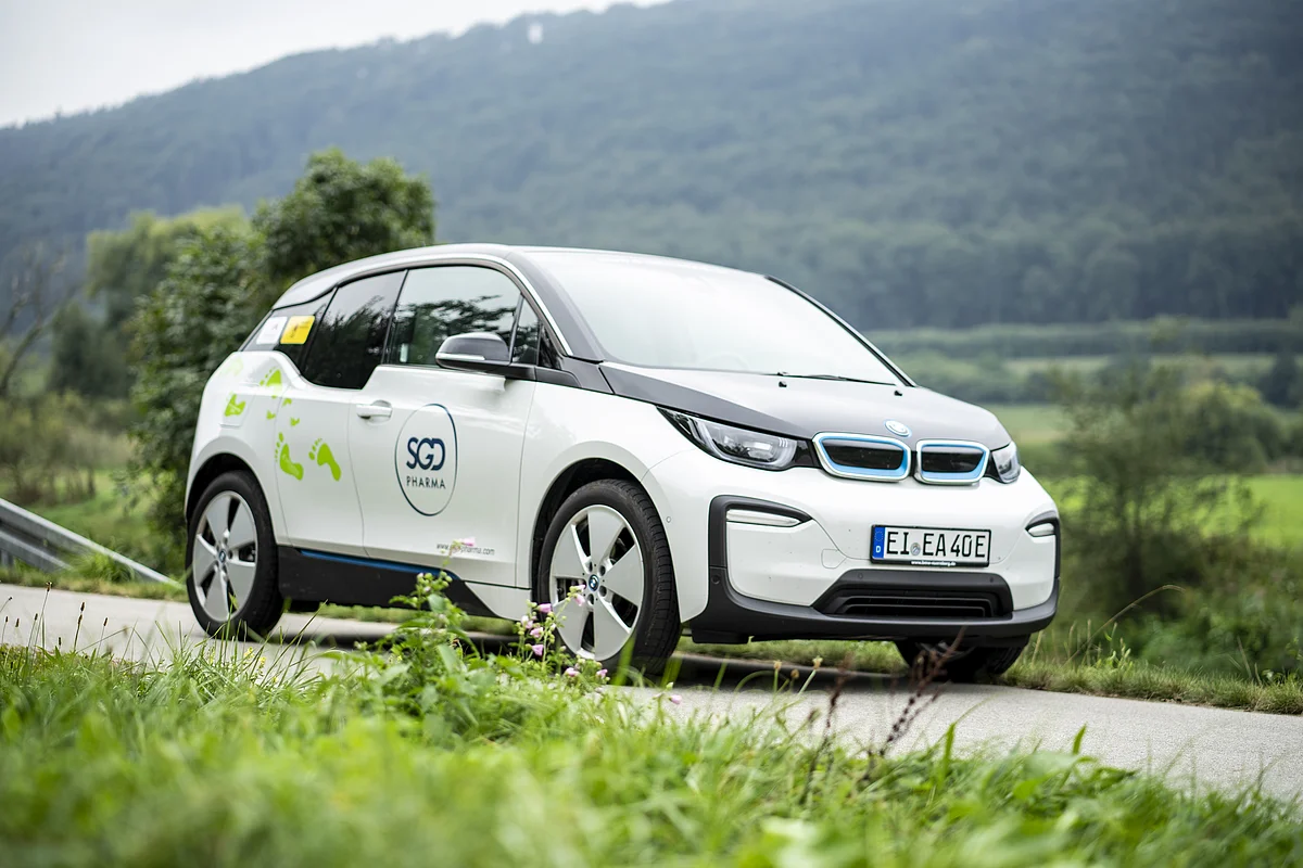 Ein weißes Elektroauto mit SGD Pharma-Logo fährt auf einer ländlichen Straße, im Hintergrund sind Bäume und Hügel.