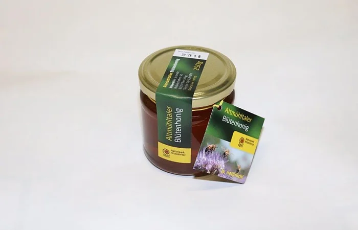 Ein Glas Altmühltaler Blütenhonig mit goldenem Deckel und grünem Etikett auf weißem Hintergrund.