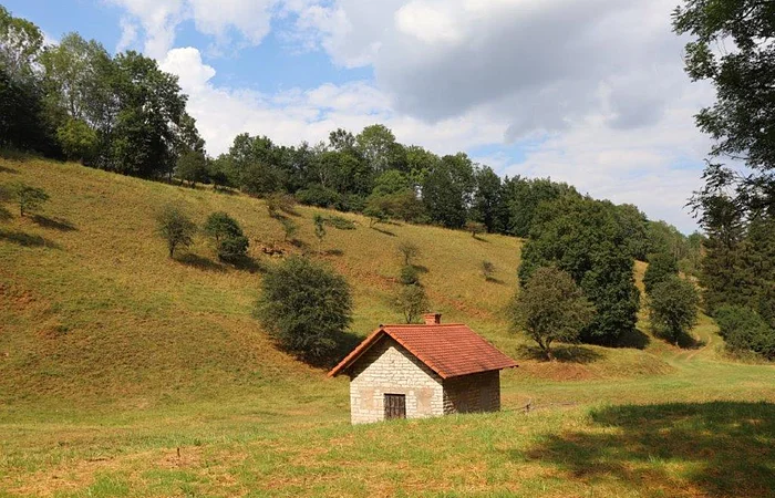 Kleines Steinhaus mit rotem Dach in hügeliger Landschaft, umgeben von Bäumen und Wiesen.