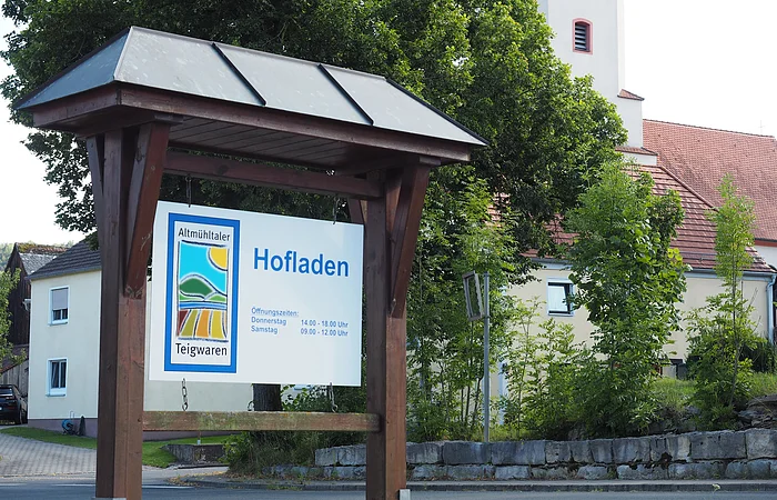 Hofladen-Schild mit Öffnungszeiten, im Hintergrund ein Gebäude mit rotem Dach und Bäumen.