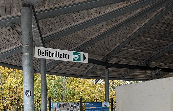 Überdachter Rastplatz mit Holzbänken, Infotafeln und einem Defibrillator an der Wand.