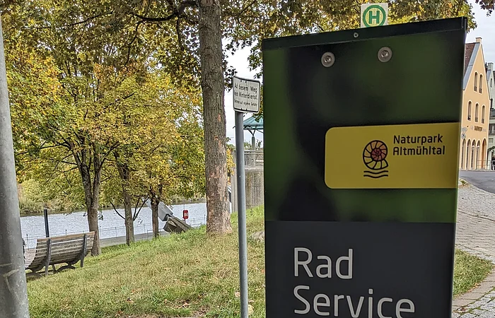 Fahrradservicestation mit Werkzeug- und Fahrradsymbol, im Hintergrund Bäume und ein Fluss.