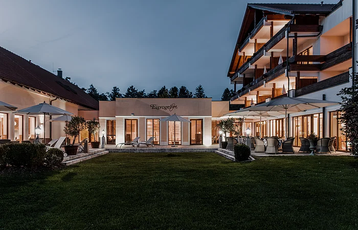 Außenansicht eines beleuchteten Gebäudekomplexes mit Terrasse und Garten bei Dämmerung.