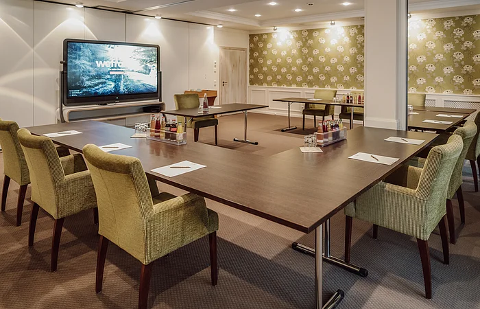 Konferenzraum mit U-förmigem Tisch, grünen Stühlen, einem großen Bildschirm und Getränken auf dem Tisch.