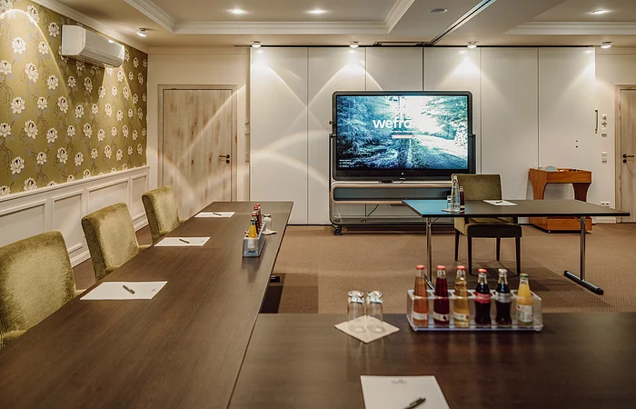 Konferenzraum mit Tischen, Stühlen, Getränken und einem großen Bildschirm an der Wand.