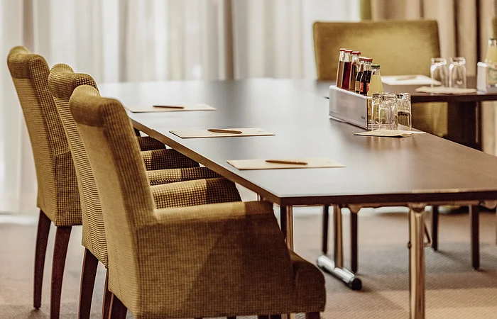 Konferenztisch mit Stühlen, Schreibunterlagen und Stiften. Auf dem Tisch stehen Getränke und Gläser.