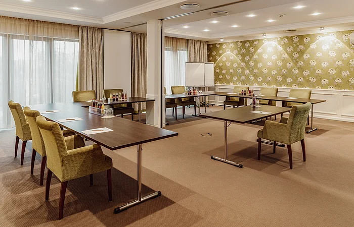 Konferenzraum mit Tischen in U-Form, grünen Stühlen, Flipchart und Getränken auf den Tischen.